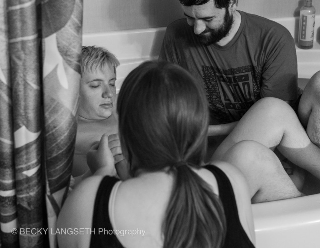 A mother labors in her bathtub for her home birth in Seattle as her husband and doula support her. This image is another reason why it's important to hire a birth photographer, because it captures the progress of birth.