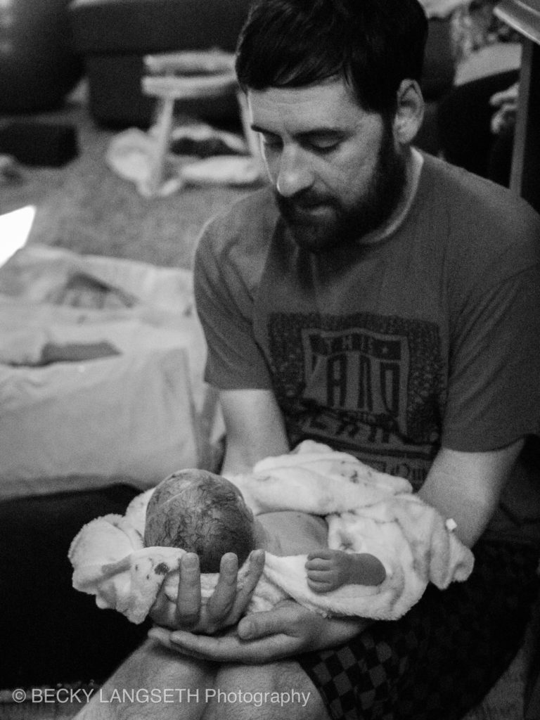 A brand new father holds his newly born baby on his lap as he gazes down at the new infant after a home birth in Seattle, Washington. This image shows the value of hiring a birth photographer to capture this special moment.