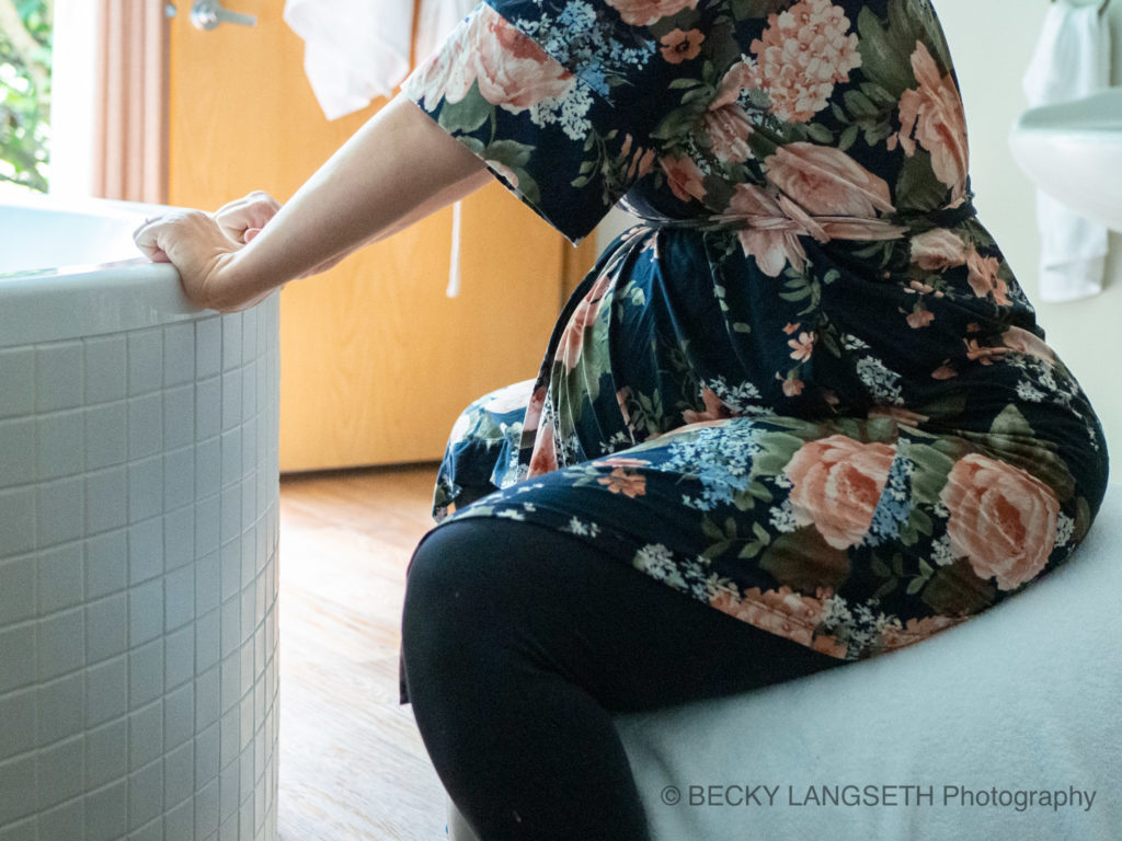 A pregnant women is leaning up against a birthing tub as she labors in a floral robe at a birth center in Seattle, Washington. Her robe is an example of what to wear during labor for great birth photos by Becky Langseth.