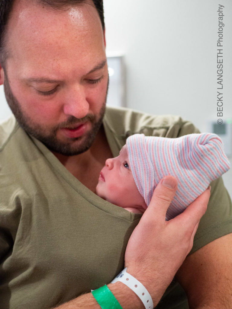 A brand new dad is holding his infant in his shirt giving the baby some skin to skin time after at birth at EvergreenHealth Family Maternity Center in Kirkland, Washington. Since Dad is wearing a plain shirt, this image is an example of what Dad's should wear to a birth by Becky Langseth Photography.