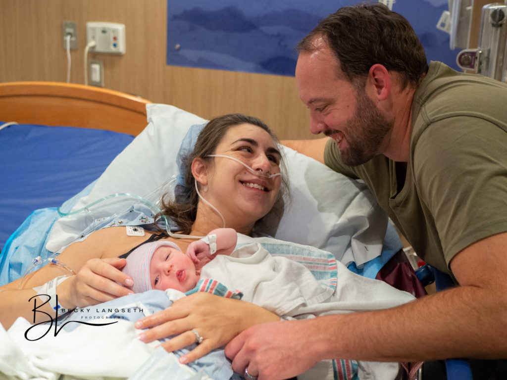 A new mother laying in a hospital bed at EvergreenHealth in Kirkland, WA looks at her husband, who just became a new dad. The two new parents smile at each other as the baby on the mother's chest looks at the camera.