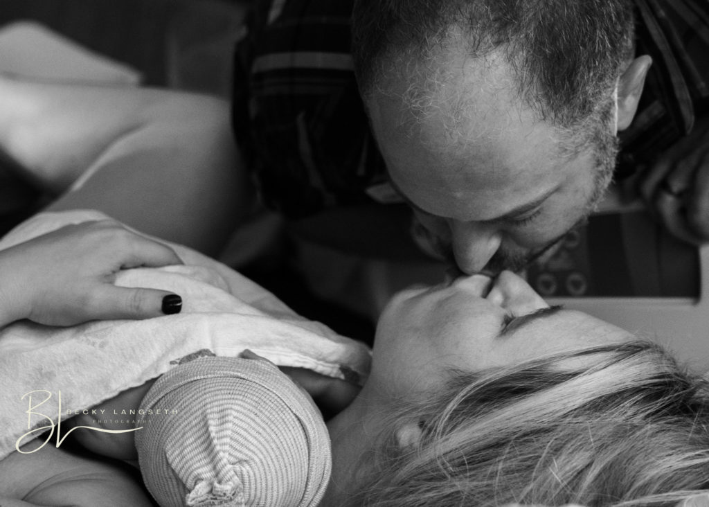A brand new dad leans over a hospital bed to kiss his wife who is holding their newly born baby at Overlake Childbirth Center. This beautiful image is an example of overcoming honest misconceptions about birth photography and shows the emotions and power of birth photography, captured by Becky Langseth, Seattle birth photographer.