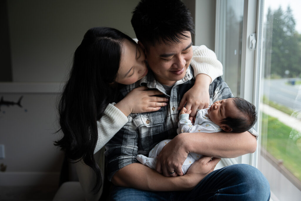 Seattle Family Photographer couple snuggling next to the window as they hold their newborn baby