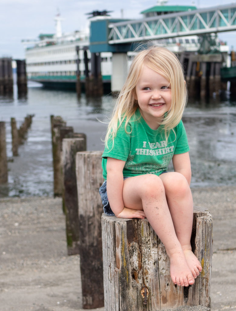  a little girl is sitting on Edmonds beach smiling slyly with the ferry docked in the background, taken by Edmonds family photographer, Becky Langseth.