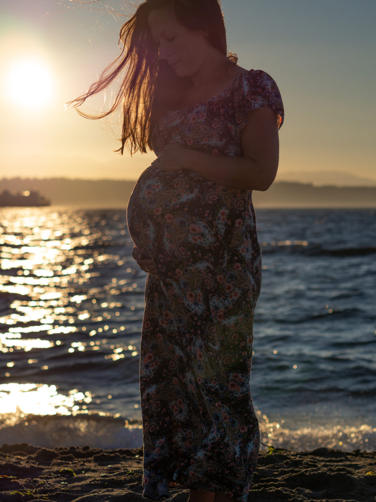 A pregnant woman is standing on the beach at golden hour with the wind blowing her hair, by Edmonds family photographer, Becky Langseth.