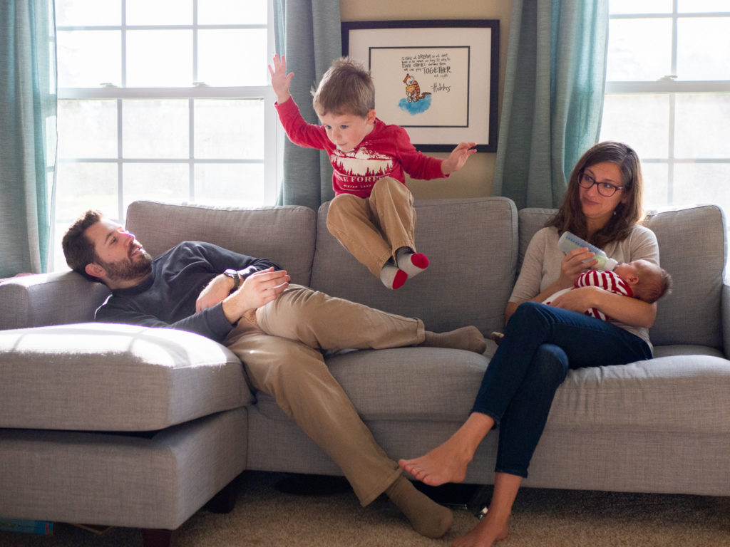 a boy jumps off the couch as his parents sit and watch during a in home family photo session in Edmonds, WA by Edmonds Family Photographer, Becky Langseth.