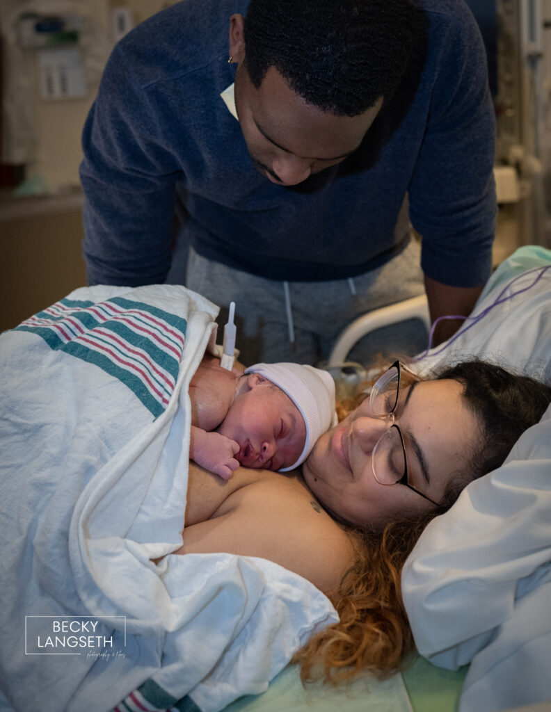 A father leans over his wife laying in a hospital bed who is holding their newly born baby at the Family Birth Center at St. Michael Medical Center in Silverdale, WA taken by a birth photographer.