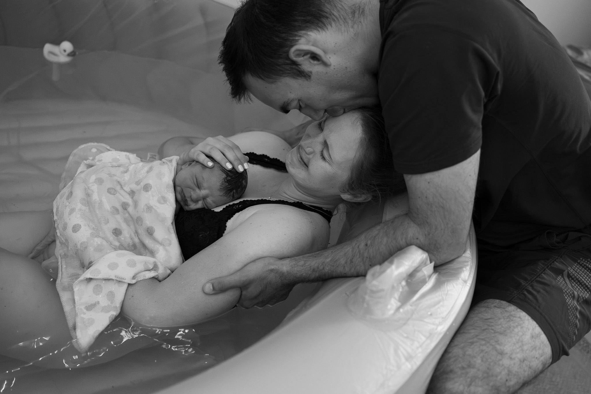 A husband is embracing his wife who just gave birth in a birth pool at home in Seattle. She is holding her newly born baby and her face is emotional.