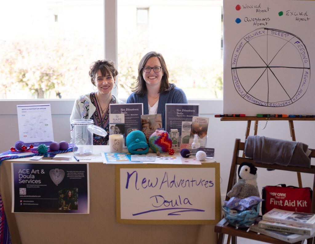 A birth doula and postpartum doula smile as they sit behind their vendor table with information for pregnant people on doula services in Seattle, WA.