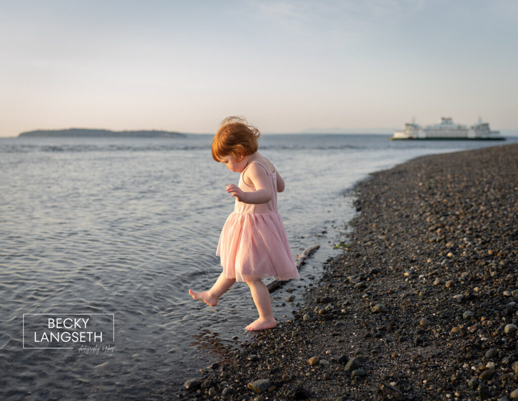 A little girl steps into the water at Mukilteo Lighthouse Beach Park. There is a ferry in the distance.