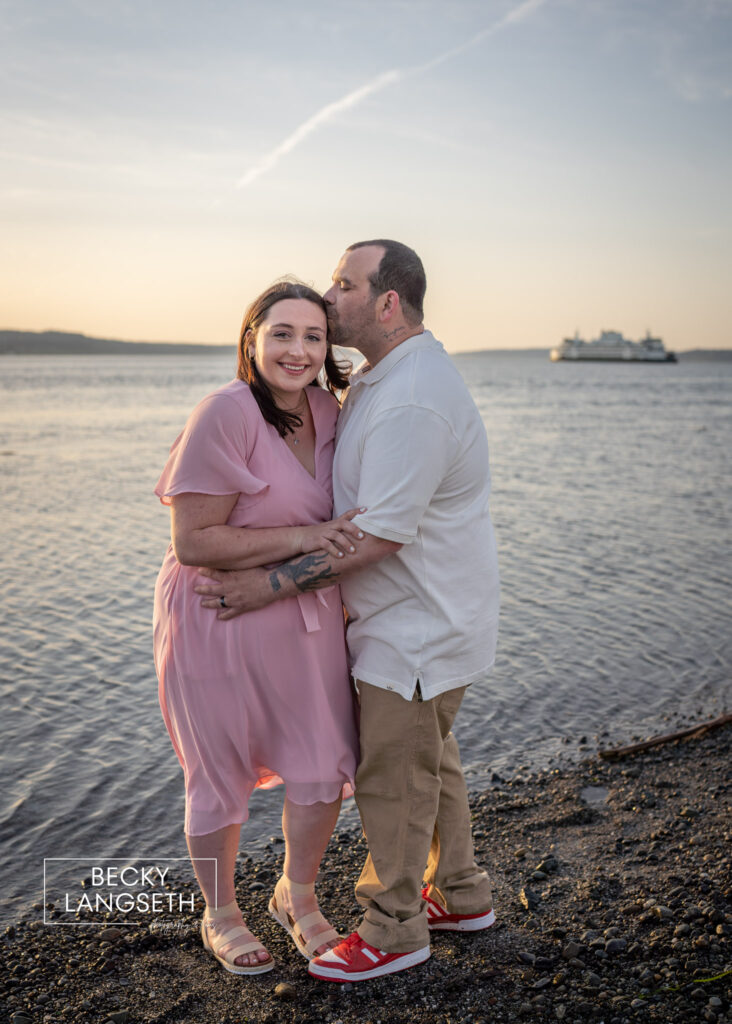 A couple stand at the beach at sunset with a Washington State ferry behind them in the far distance. The man is kissing his pregnant partner's forehead as she smiles.