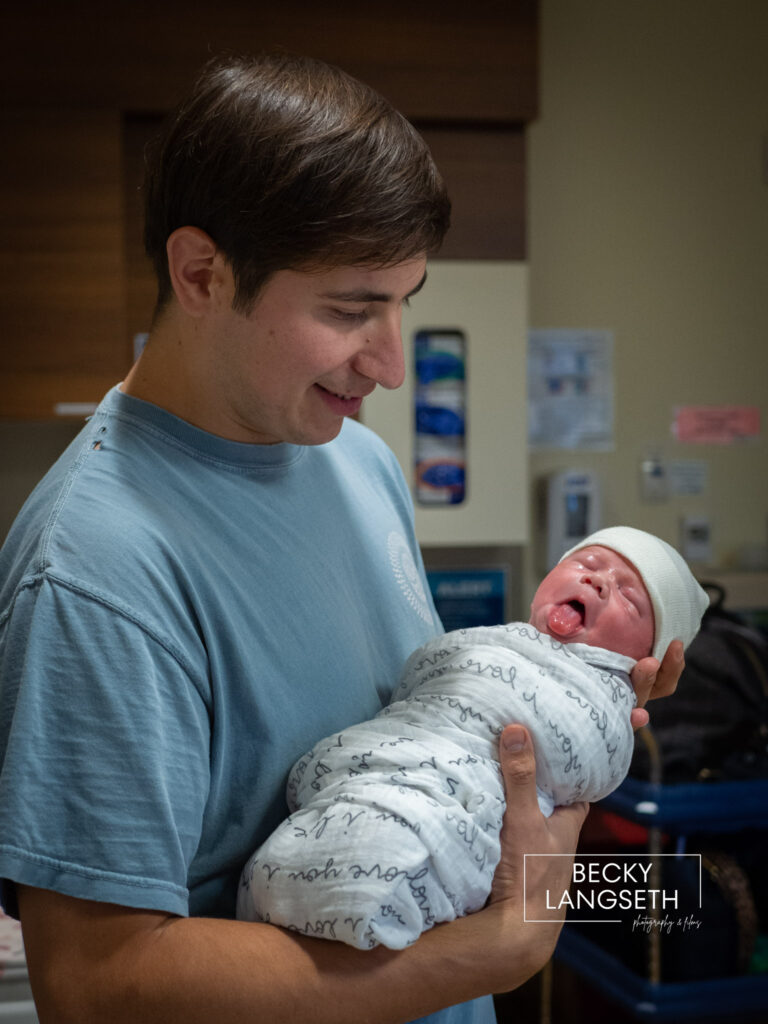 A new dad holding his infant son for the first time at Swedish Issaquah Hospital. The baby is sticking out it's tongue. 