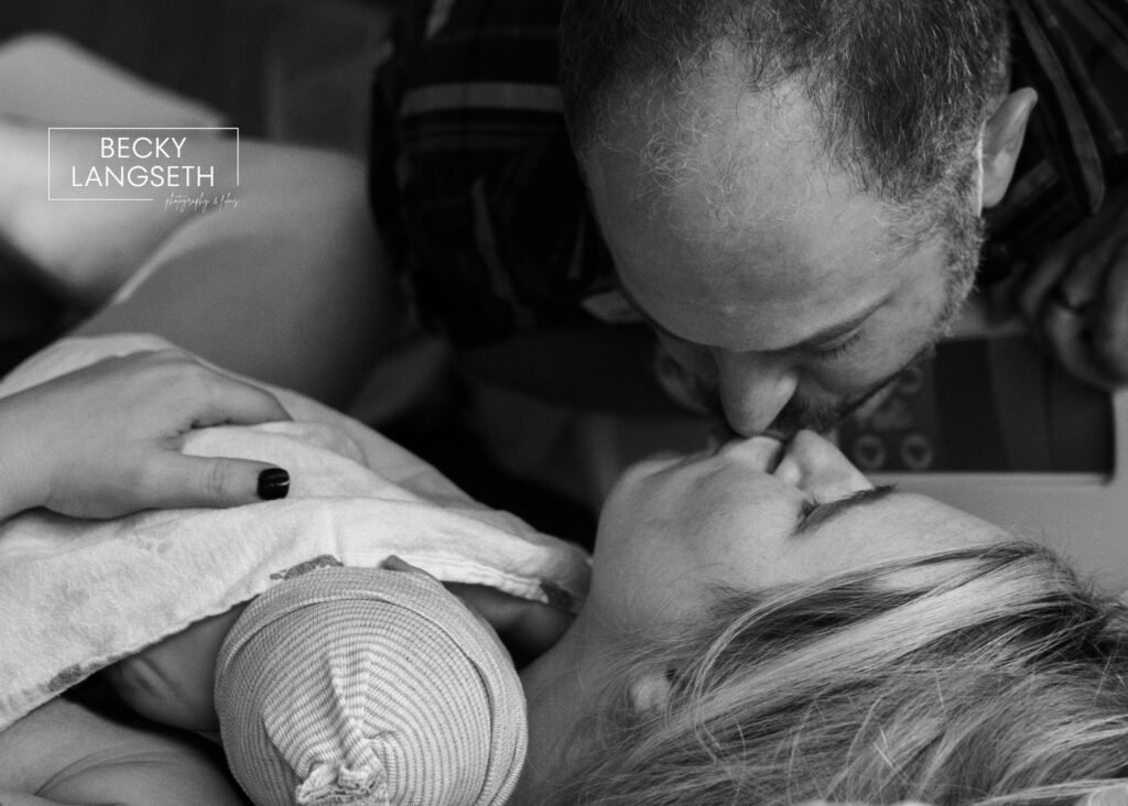 A husband and wife kiss after the birth of their new baby as the mother is lying on the hospital bed with their baby in her arms. 