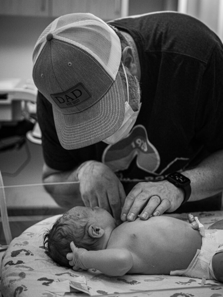 A new dad wearing a "dad" hat looks at his newly born son in the baby warmer in a hospital room. 