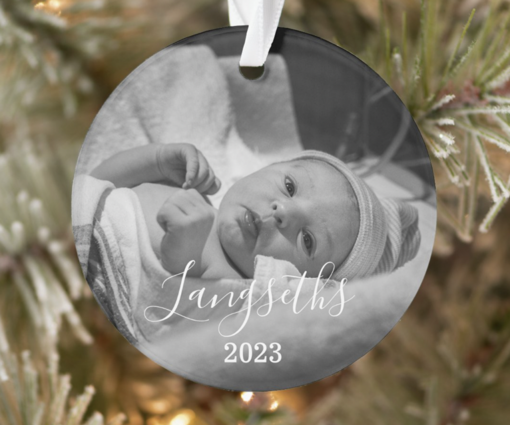 A round holiday ornament hanging from a tree with a photo of a newborn baby at the hospital.