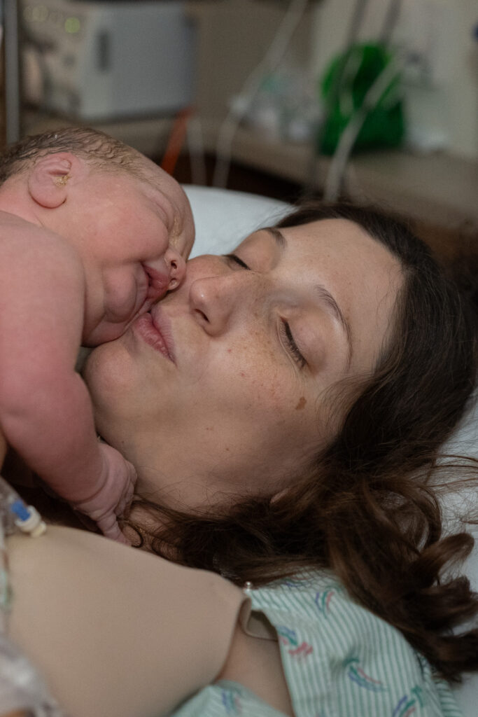 a mother holds her baby up to her face check-to-cheek in a hospital bed after baby has recently been born