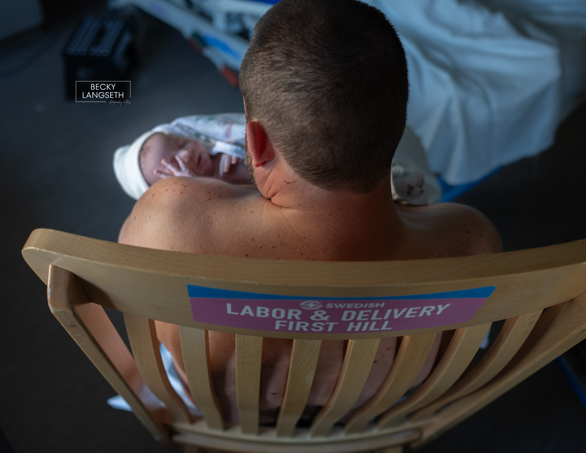 A father hold his newborn baby skin to skin in a rocking chair at Swedish First Hill Birth Center with the hospital name on the back of the rocking chair.