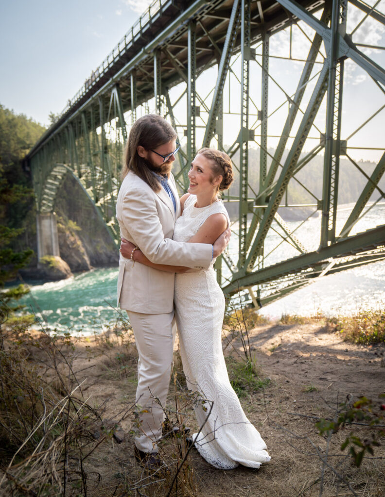 Bride and groom embracing and looking at each other with the Deception Pass bridge in the background.