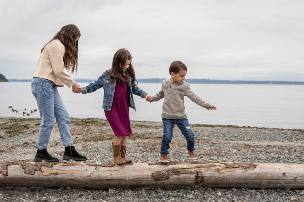 Three kids are holding hands and trying to balance as they walk along a driftwood log at Carkeek Park beach