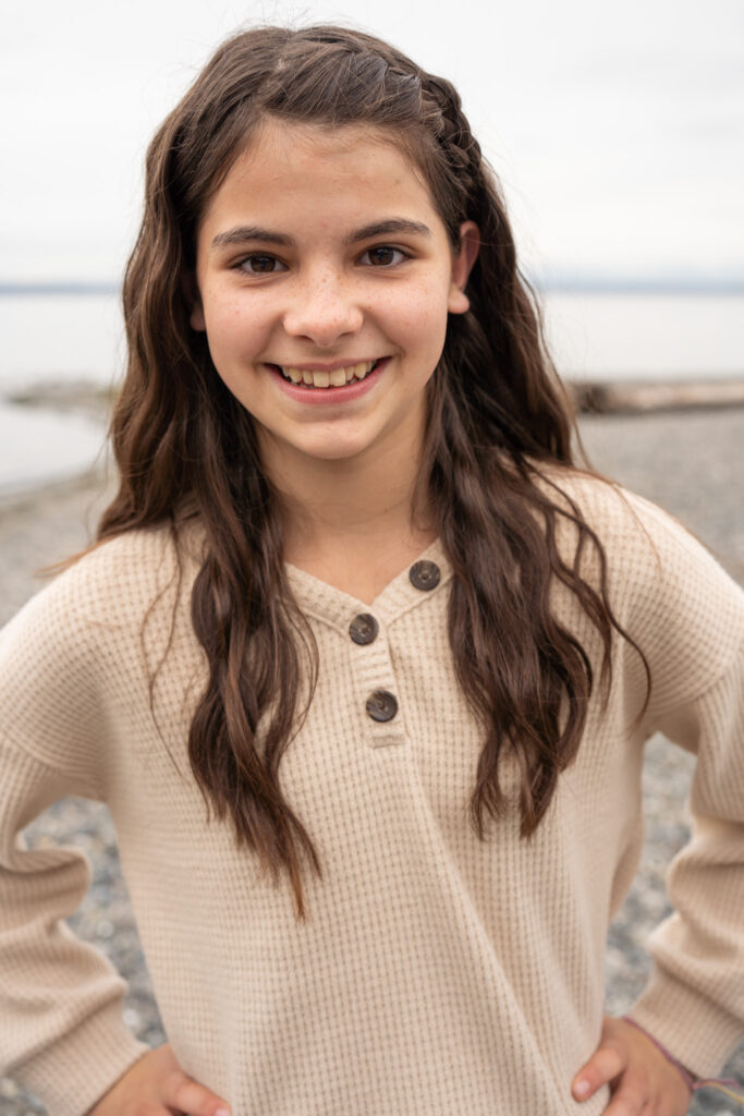 a pre-teen girl grins at the camera with the beach in the background on a dreary PNW day.