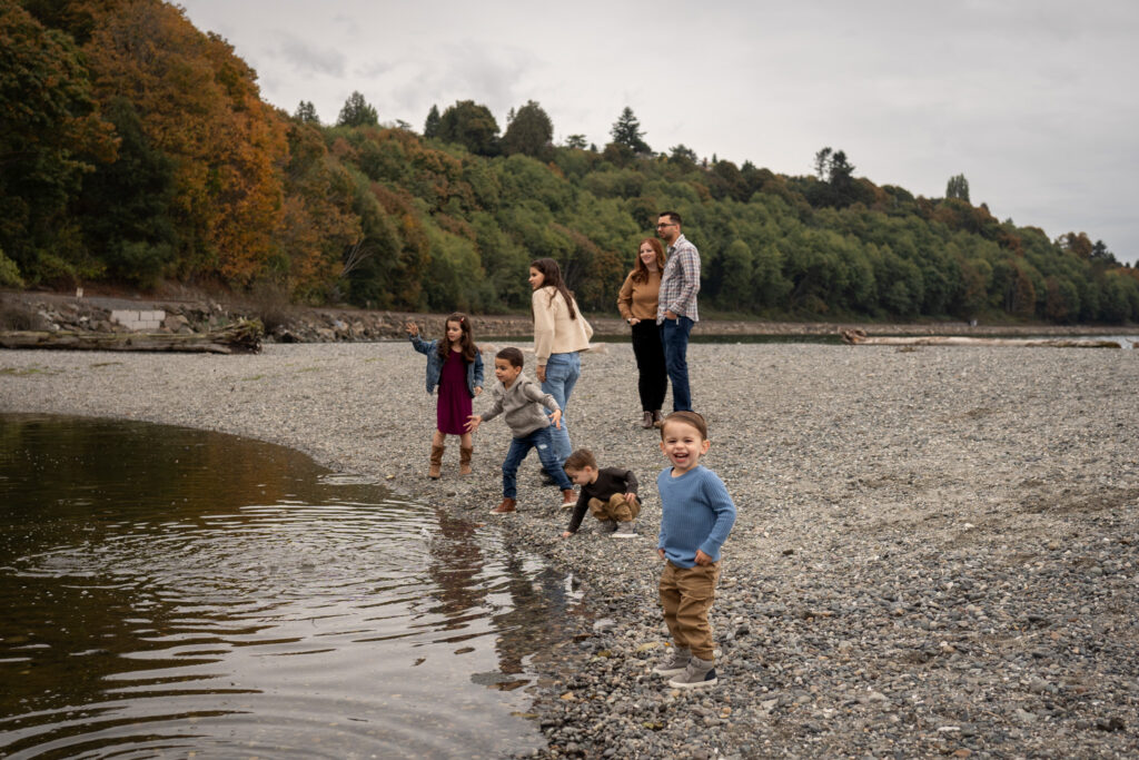 Mom and dad are standing in the background as their 5 kids are trying to skip rocks on an inlet at Carkeek Park beach
