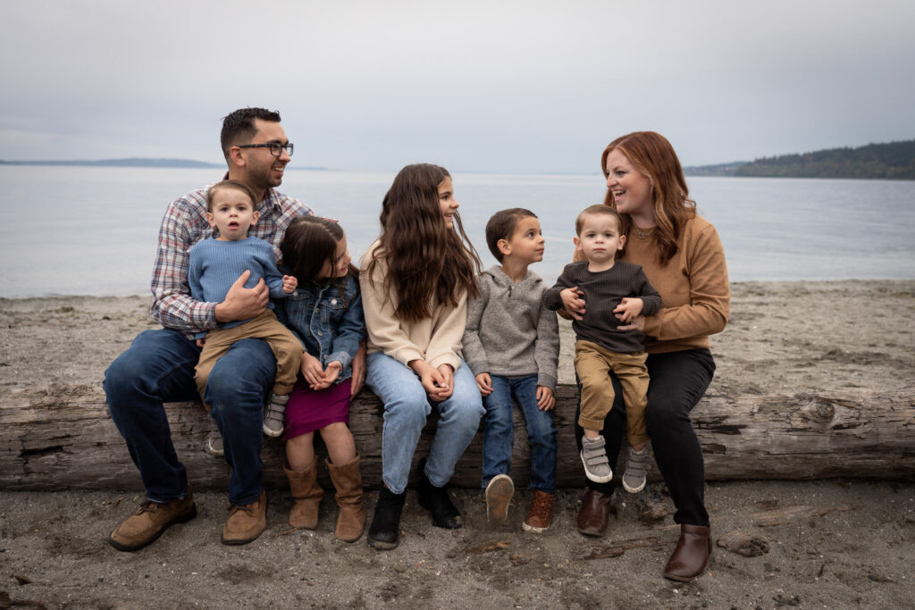 Family of 7 is sitting on a log at Carkeek Park on the beach with the Puget Sound in the background.