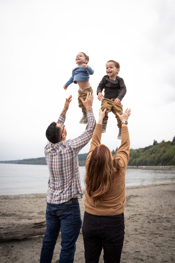 A mother and father are tossing their twin toddlers up in the air at Carkeek Park.