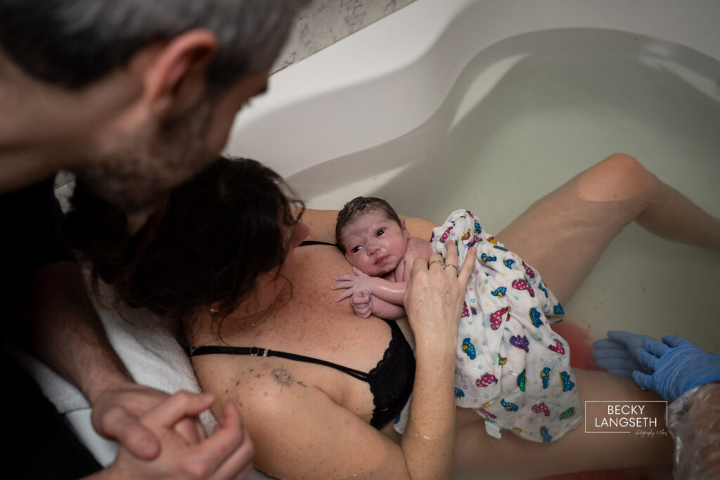 A birds-eye view of a mother in a birthing tub holding her baby after giving birth. The baby is wide-eyed and looking up at her parents at the Puget Sound Birth Center.