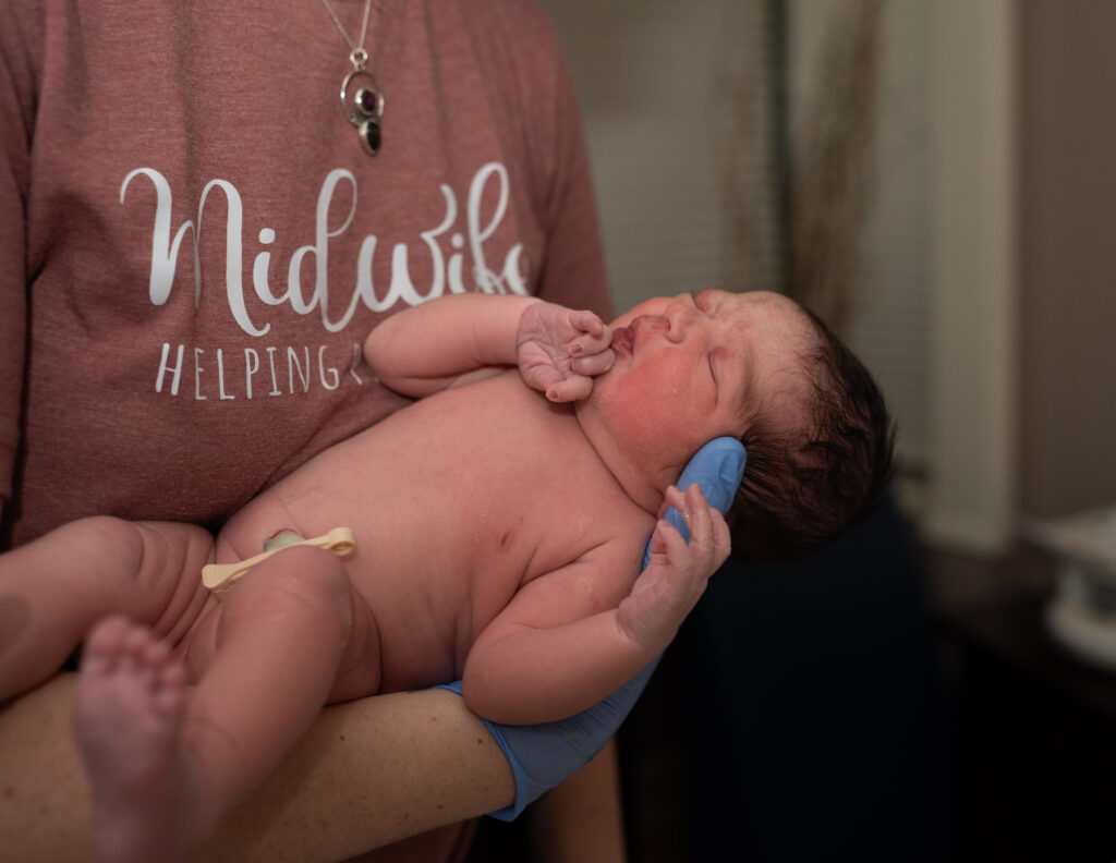 Midwife is holding baby and wearing a shirt that says, "Midwife."