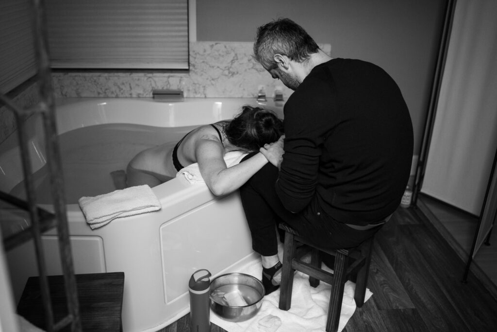A black and white image of a husband leaning over and holding his pregnant wife's hands as she continues to labor in a birthing tub