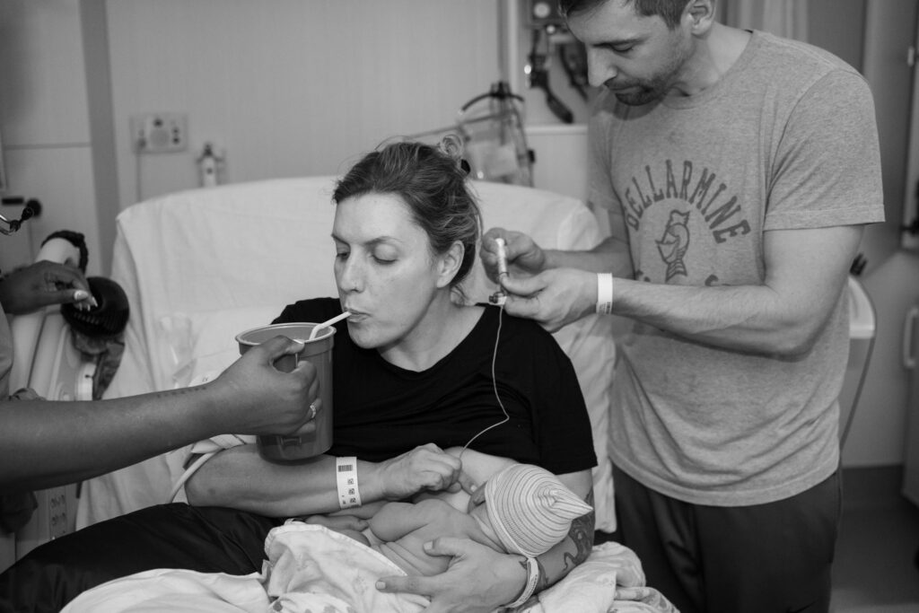 A mother takes a drink after giving birth at UW Northwest Childbirth center as her husband is setting up a feeding tube for their new baby.