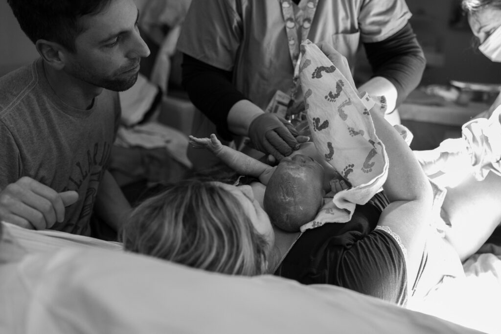 a brand newly born baby is laying tummy side up on it's mother's chest with it's arms lifted high as the father overlooks from above.