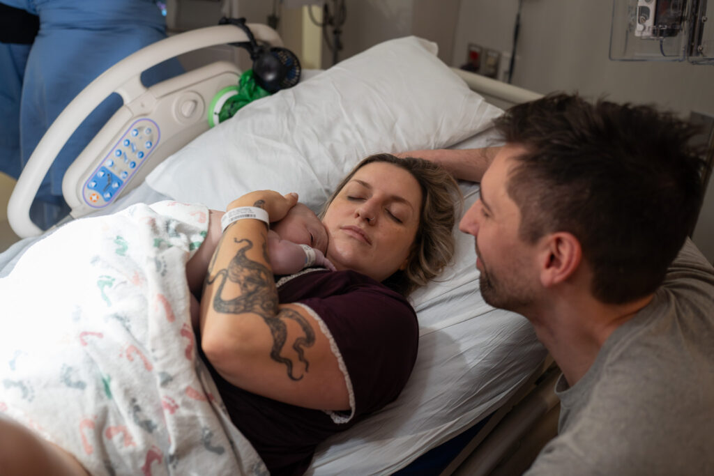 A mother snuggles her baby in relief with her eyes closed as the father smiles over her hospital bed.