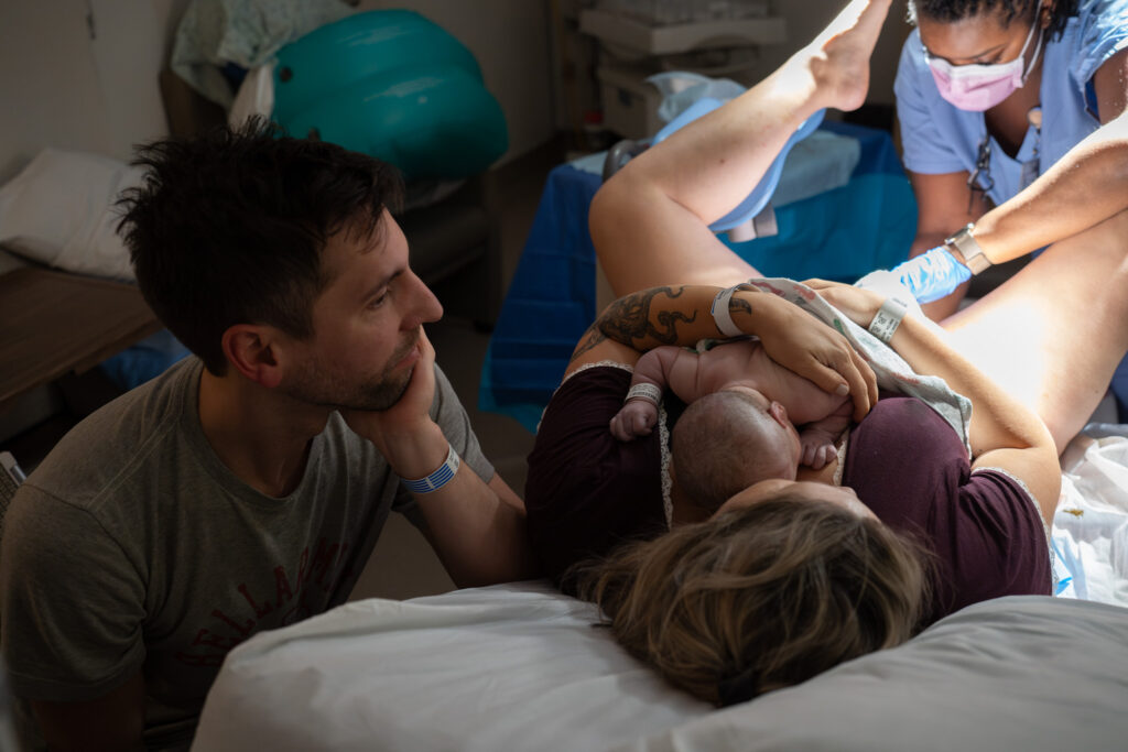 A mother is getting stitched up while laying on a hospital. bed after tearing a bit when giving birth. She is holding the baby and her husband is next to her looking at the baby. 