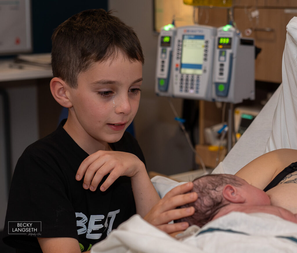 big brother has a tear rolling down his face as he meets his new sibling just an hour after being born