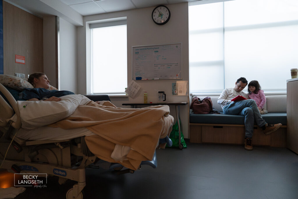 father reads to his daughter on the hospital bench as the mother lays in the hospital bed.