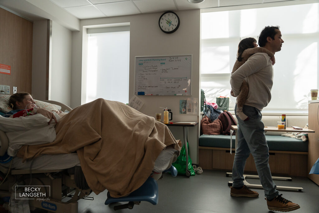 soon-to-be big sister rides on her father's back piggy back style as they wait for the new baby to be born at Evergreen Hospital