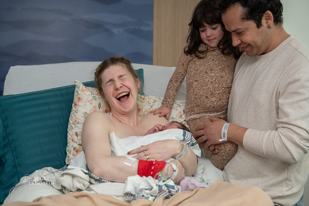 a mother laughs while holding her newly born baby as her husband holding her older daughter look at the new infant