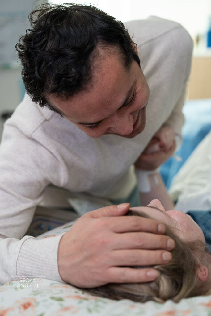 husband puts his hands on his wife's head smiling at her and holding her hand as she is about to give birth