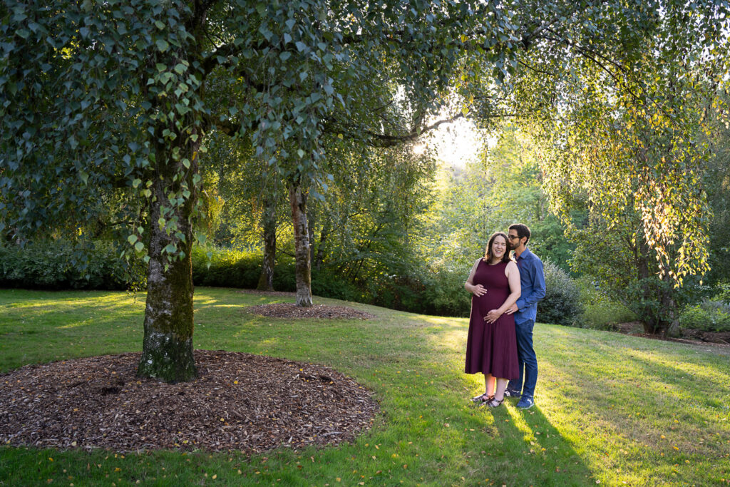 A couple standing at a park. The woman is holding her pregnant belly, and her husband kisses her forehead.