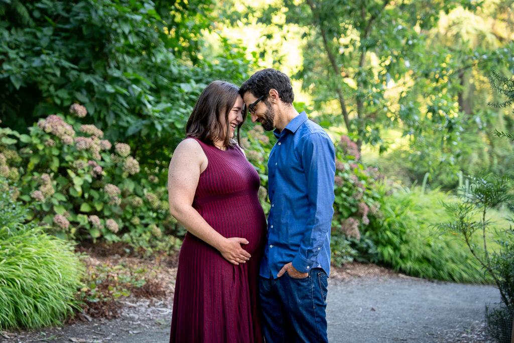 a pregnant couple laugh together in the lush area of a beautiful park in Seattle