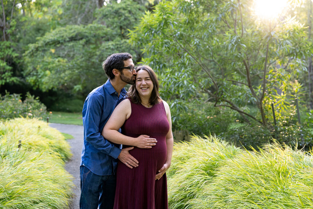 A close up backlit image of a pregnant couple with the husband kissing his wife's forehead while she is smiling at the camera and holding her hand on her pregnant belly